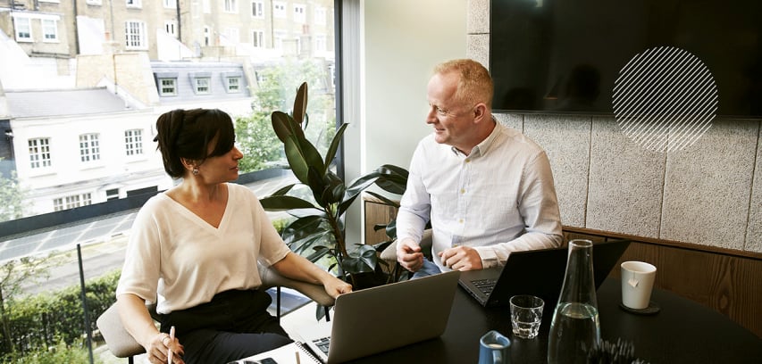 Two professionals sit in a conference room conversing in front of a large window